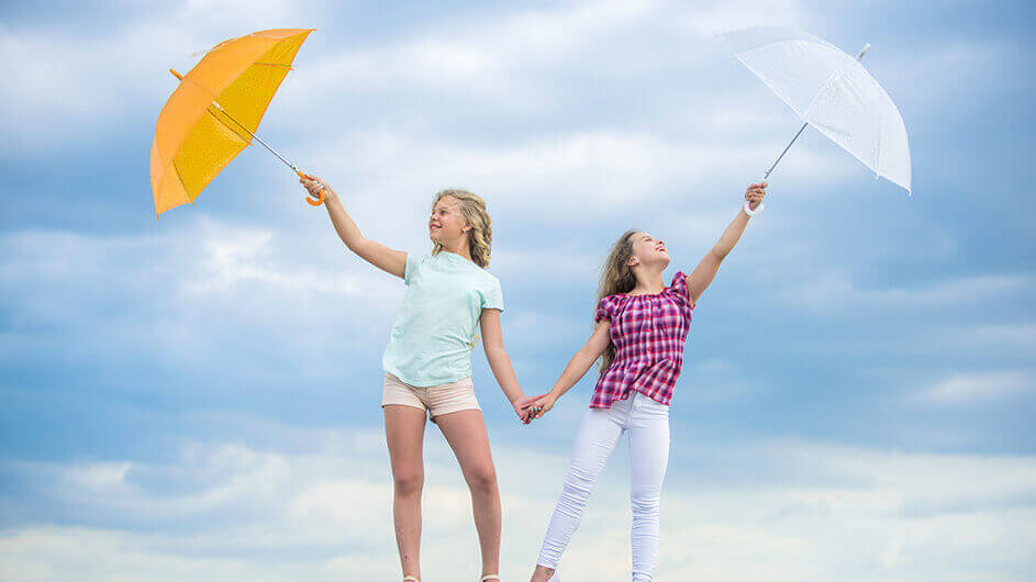 Zwei Mädchen stehen lächelnd mit Regenschirmen vorm wolkenverhangenen Himmel