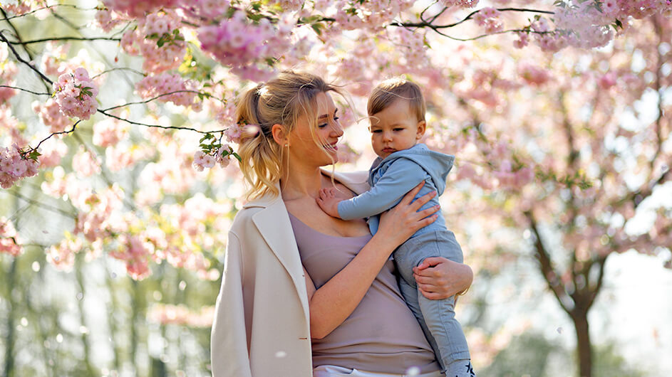Eine lächelnde Mutter steht, ihr Baby auf dem Arm haltend, unter Kirschblüten