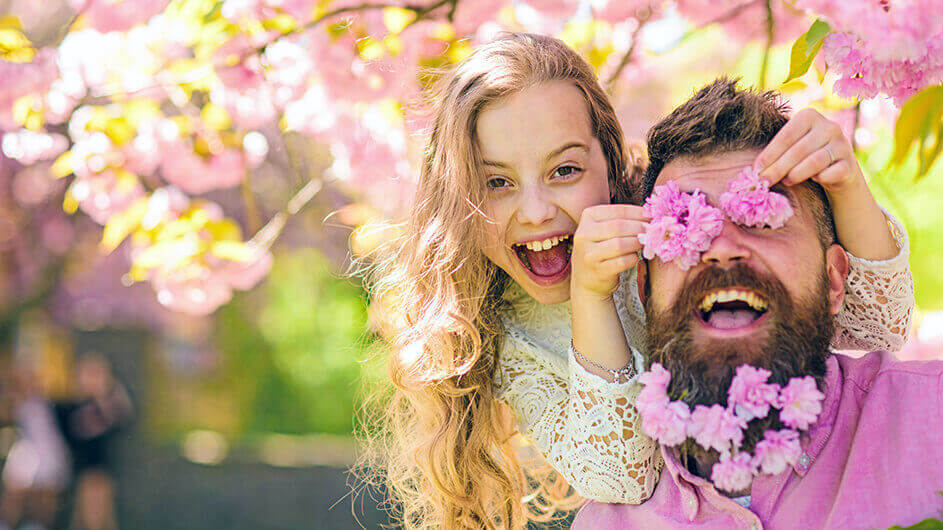 Eine lachende Tochter hält ihrem Vater Kirschblüten vor die Augen und hat auch den Vollbart ihres Vaters mit Blüten verziert.