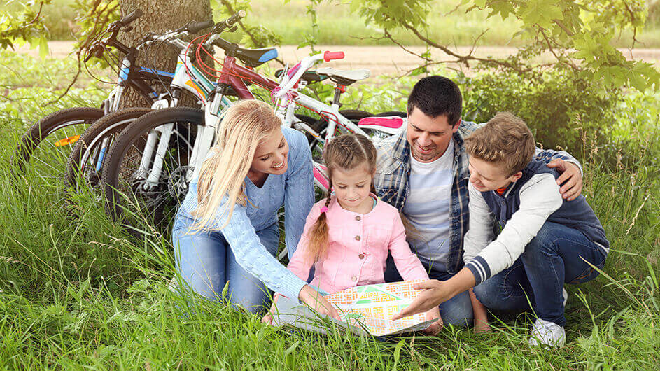 Eine Familie ist auf Fahrradtour im Grünen und guckt lächelnd auf eine Landkarte. Hinter ihnen stehen ihre Fahrräder.