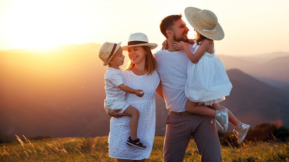 Eine glücklich lächelnde Familie vor einem Sonnenuntergang im Sommer in heller Sommerkleidung mit Strohhüten