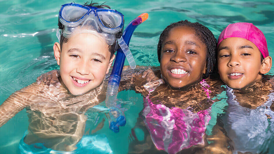 Drei Kinder im Schwimmbecken eines Freibads. Sie umarmen sich und lächeln.