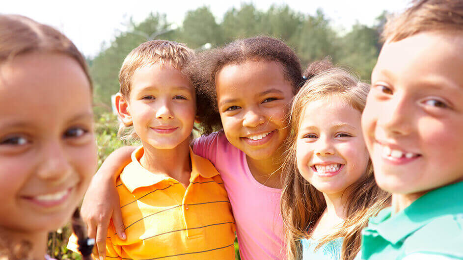 Fünf Kinder lächeln bei Sonnenschein in die Kamera und umarmen sich