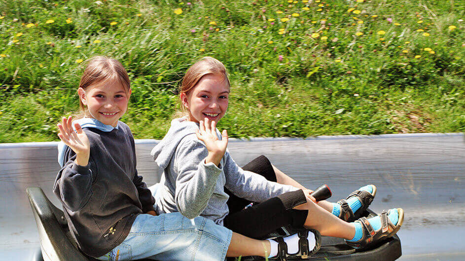 Zwei winkende und fröhlich lächelnde Kinder auf einer Sommerrodelbahn