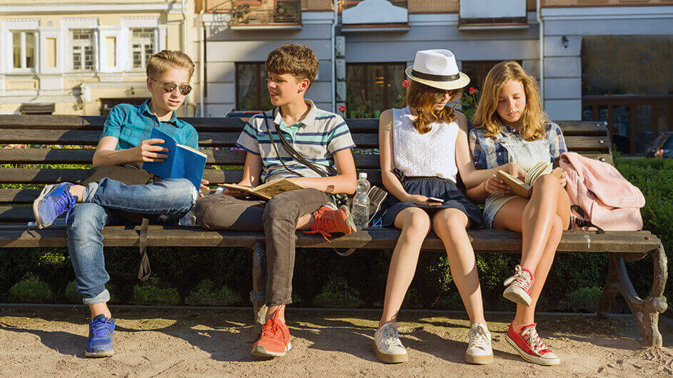 Kinder auf einer Sprachreise: Sie sitzen auf zwei Bänken, unterhalten sich und lesen in Büchern.