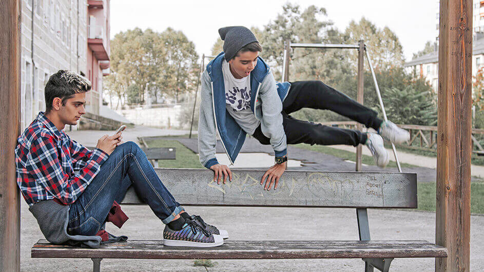 Ein Junge beim Parour-Laufen. Er springt über eine bank, während ein anderer Teenager dort mit Smartphone sitzt.