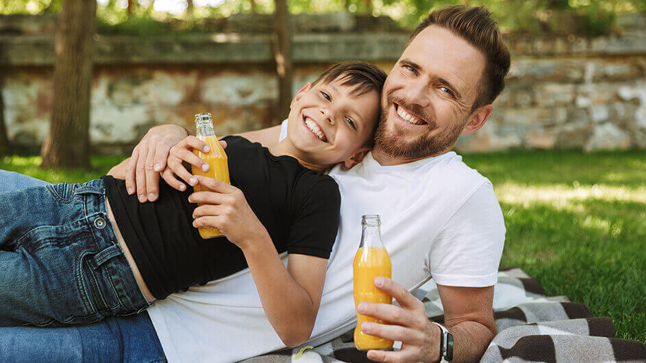 Ein Sohn liegt angelehnt an seinen Vater im Park. Beide trinken Saft aus Glasflaschen und lächeln fröhlich in die Kamera.