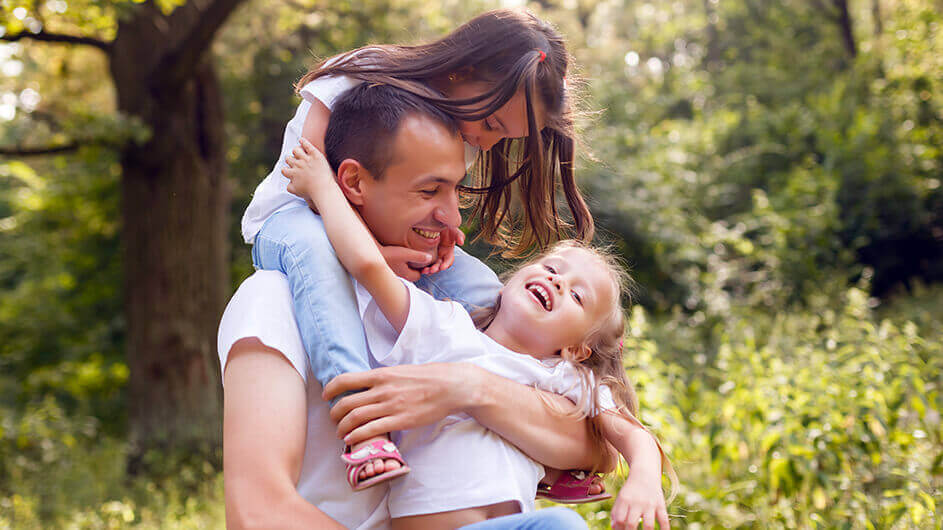 Ein Vater spielt mit seinen zwei kleinen Töchtern im Park. Die eine Tochter sitzt auf den Schultern, die andere hält er im Arm. Alle lachen.