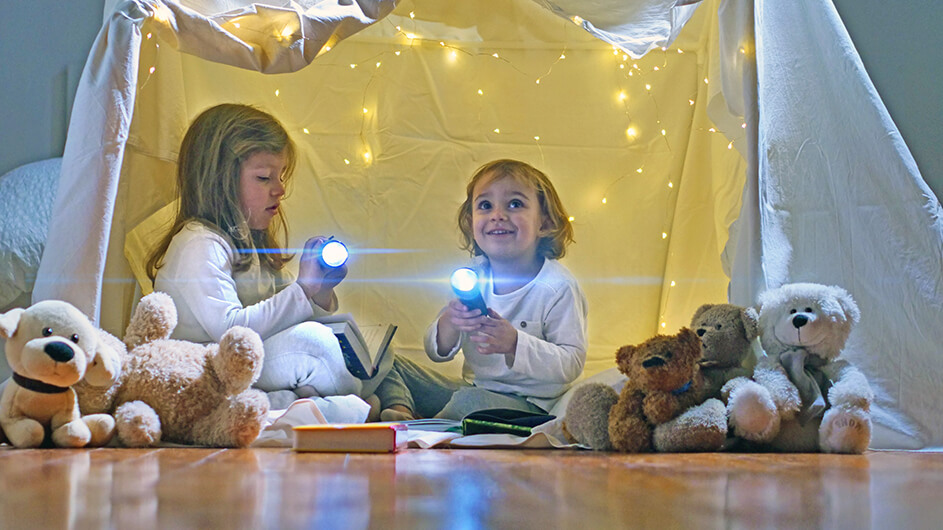 Zwei kleine Kinder in einer Höhle aus Decken mit Taschenlampen und Kuscheltieren